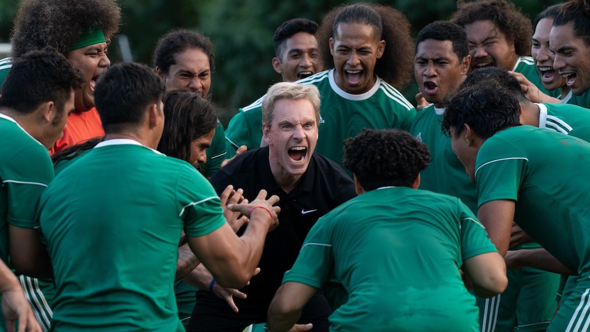 Michael Fassbender as a blonde-haired coach screaming in the middle of his soccer team players all wearing green