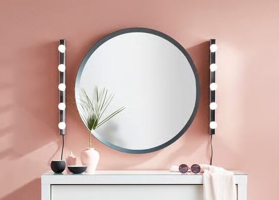 A large round grey-framed mirror with two vertical strips of round bulb wall lights mounted either side, styled above a white cabinet against a pink wall