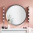 A large round grey-framed mirror with two vertical strips of round bulb wall lights mounted either side, styled above a white cabinet against a pink wall