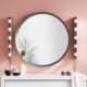 A large round grey-framed mirror with two vertical strips of round bulb wall lights mounted either side, styled above a white cabinet against a pink wall