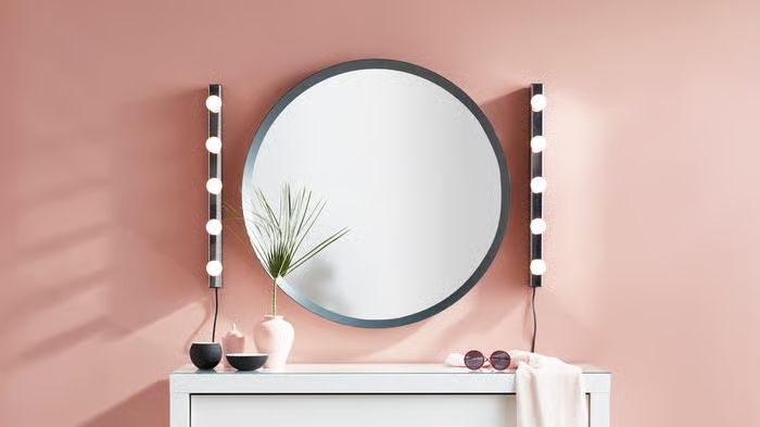 A large round grey-framed mirror with two vertical strips of round bulb wall lights mounted either side, styled above a white cabinet against a pink wall