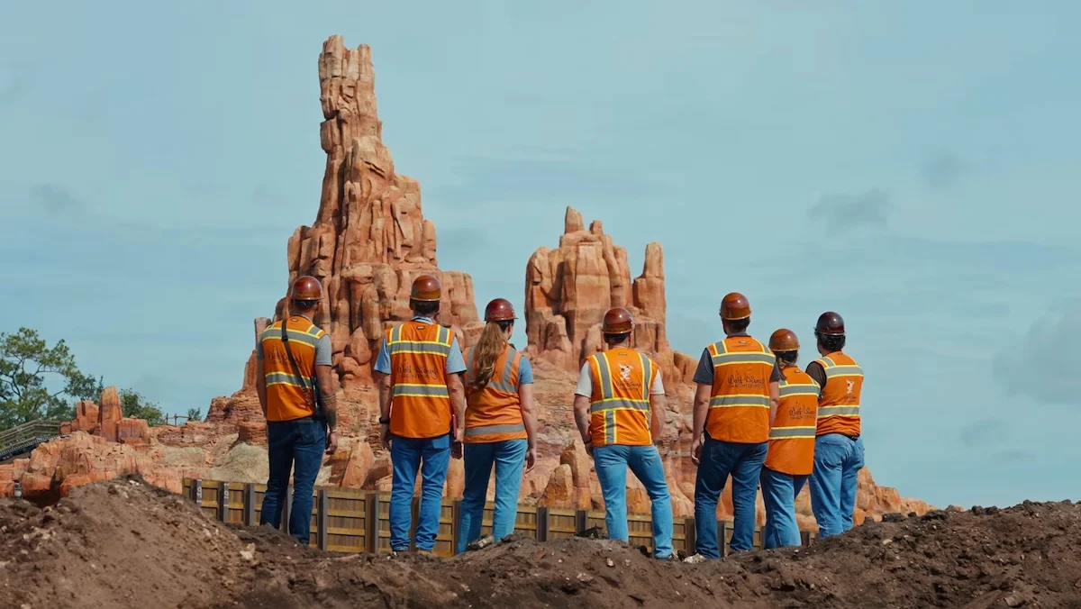 Imagineers seen from behind look at Big Thunder Mountain while wearing their orange vests and hard hats from the Imagineering Story 2